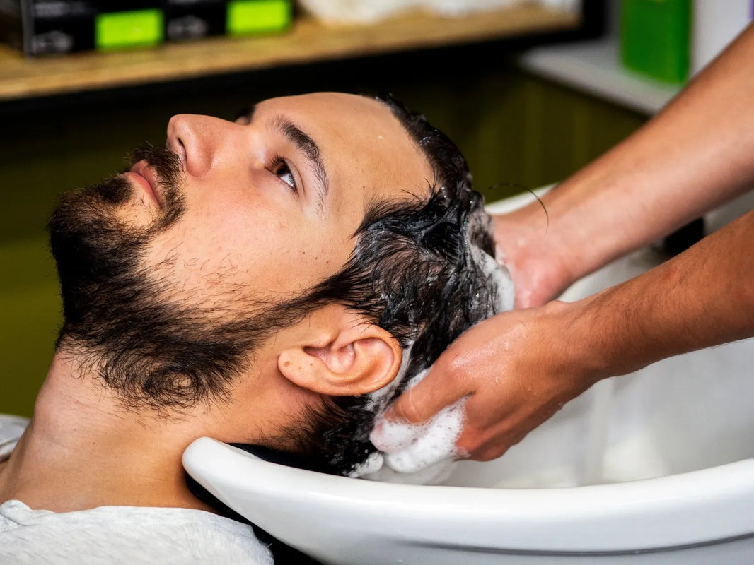 Homme barbu allongé au bac pendant un shampoing en salon, geste doux pour limiter la chute de cheveux au lavage