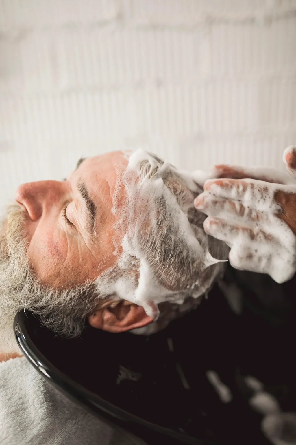 Un coiffeur masse le cuir chevelu d’un homme âgé avec un shampoing moussant pendant une routine capillaire pour stimuler la pousse des cheveux